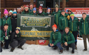Members of Clarkson's alpine ski team pose for a photo with a banner featuring Clarkson's alpine ski team logo while outside at a snowy event.
