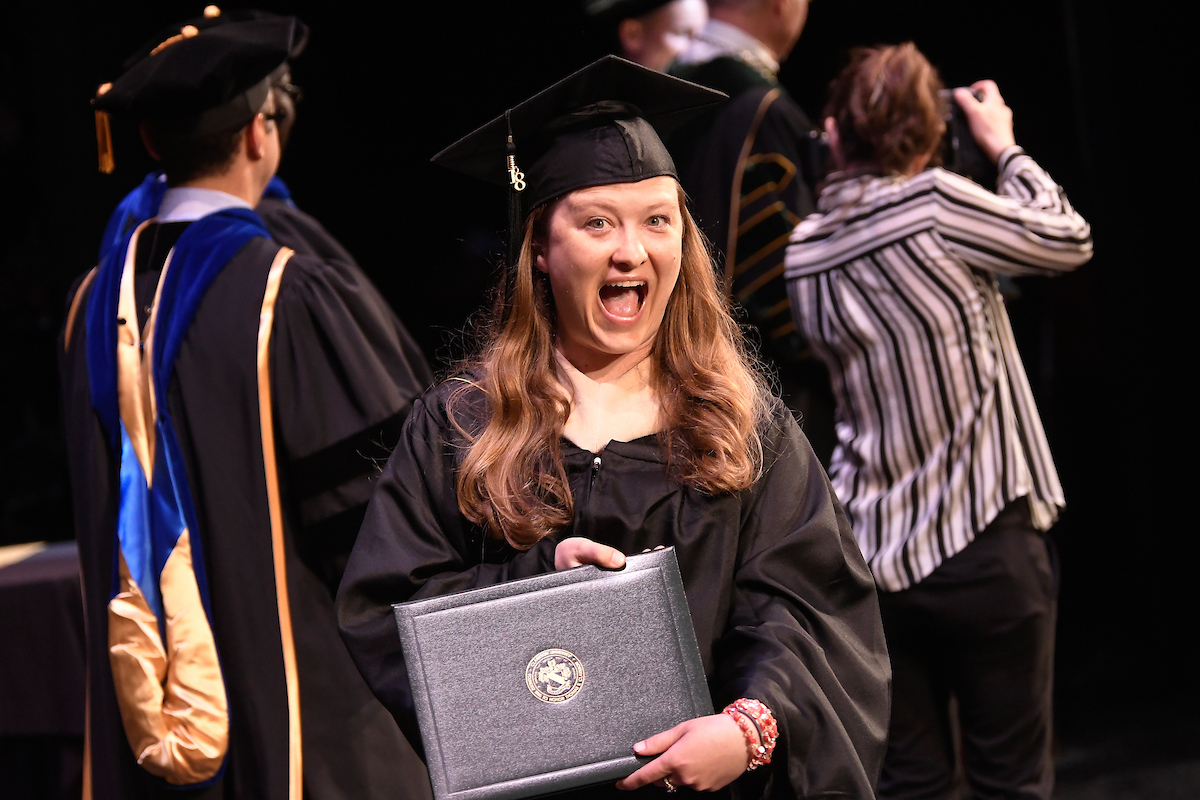 A Clarkson graduate student proudly displays her graduation diploma for the camera as she walks off stage during graduation.