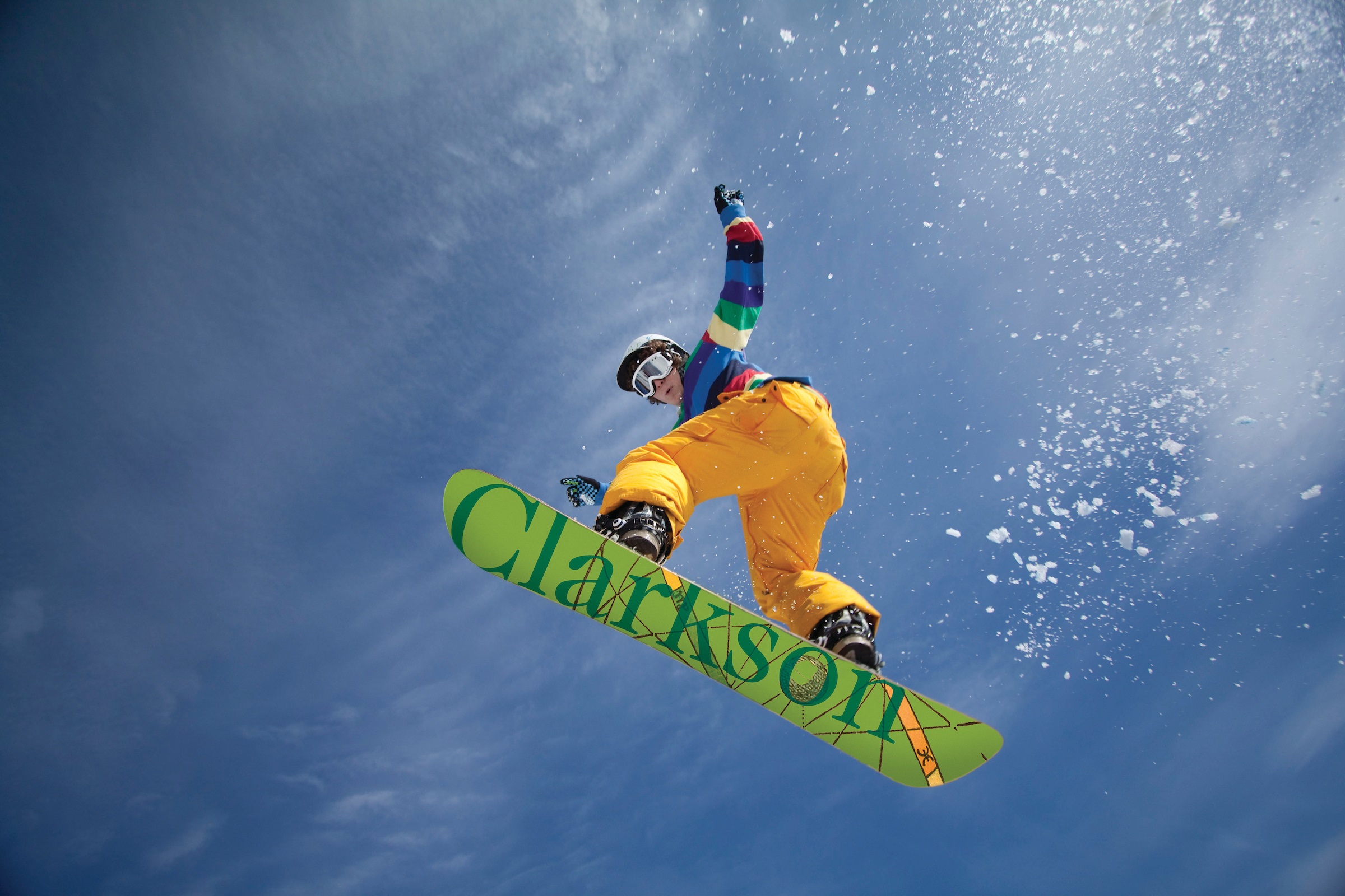 A upward shot of a snowboard and a person in yellow snow pants with text on the bottom of the snowboard that reads Clarkson