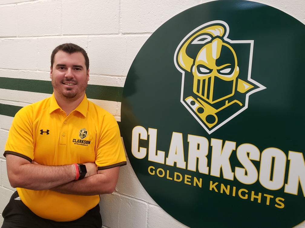 Photo of Erik Whitcombe, who oversees intramural sports at Clarkson, next to the Clarkson athletics logo on a wall.