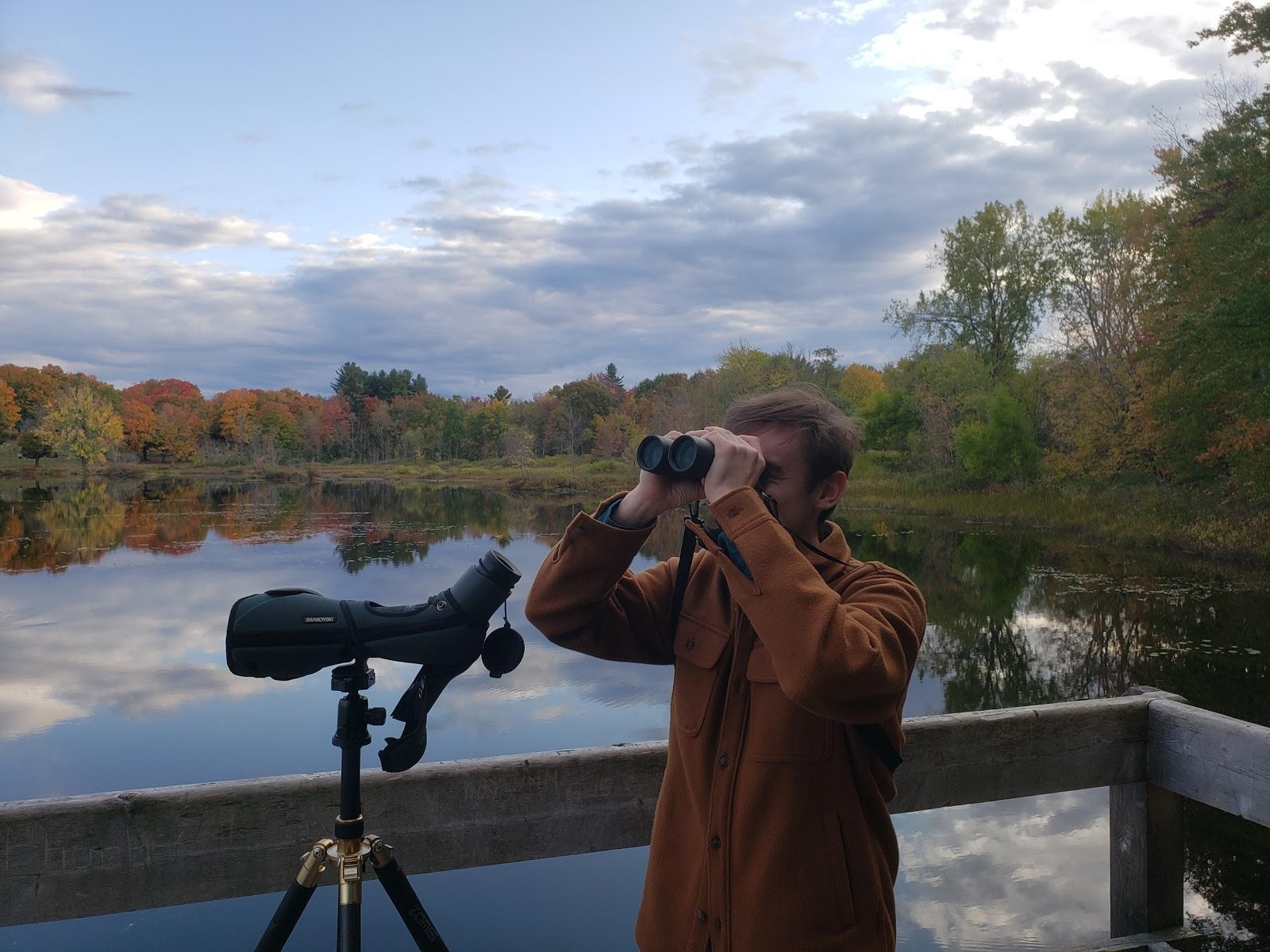 A man stands on a wooden deck using binoculars to look into the distance while a spotting scope is mounted on a tripod in front of him. In the background is a body of water, surrounded by trees.
