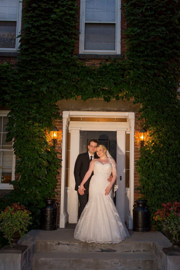 A man and woman pose in the doorway to a brick building surrounded by ivy. The woman is in a wedding dress and the man in a tuxedo. 