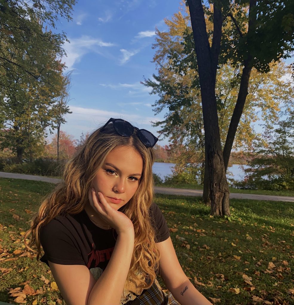 A student poses for a photo with her chin on her hand and sunglasses on her head, with blue skies and autumn leaves in the background. 