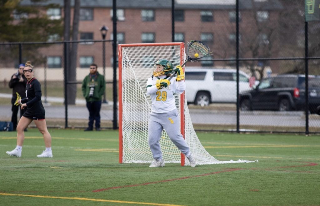 A person on the lacrosses field in full uniform standing in front of a goal holding a goalie stick with a yellow ball in the net.