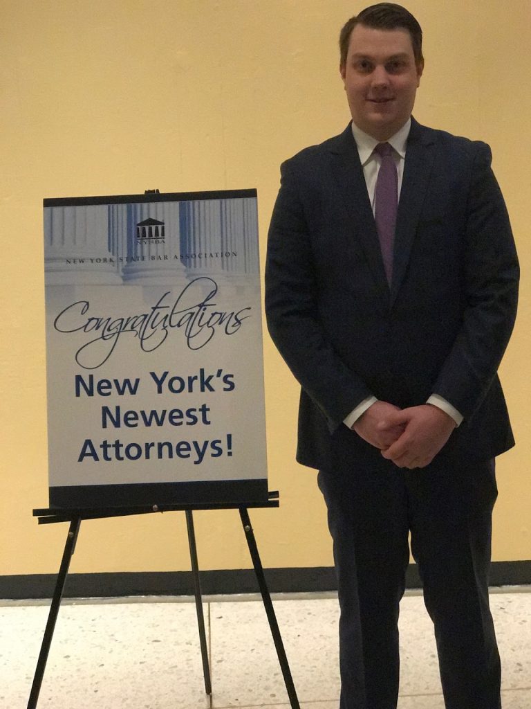 Alex Nicoles standing next to a placard displaying 'Congratulations New York's Newest Attorneys!'