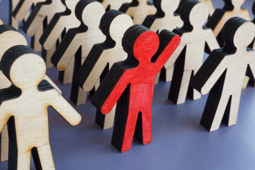 An image of a unique red person figurine standing out in a crowd of wood colored people figurines.