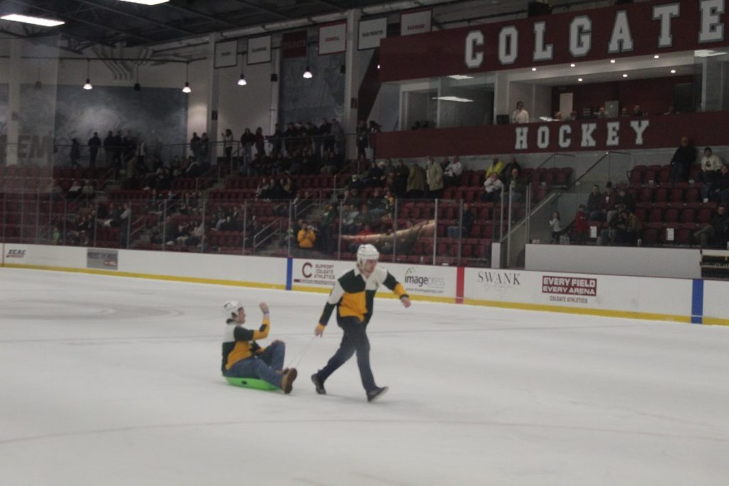 Two Clarkson Pep Band members participate in a game on the ice during an intermission. One is pulling the other on a sled.