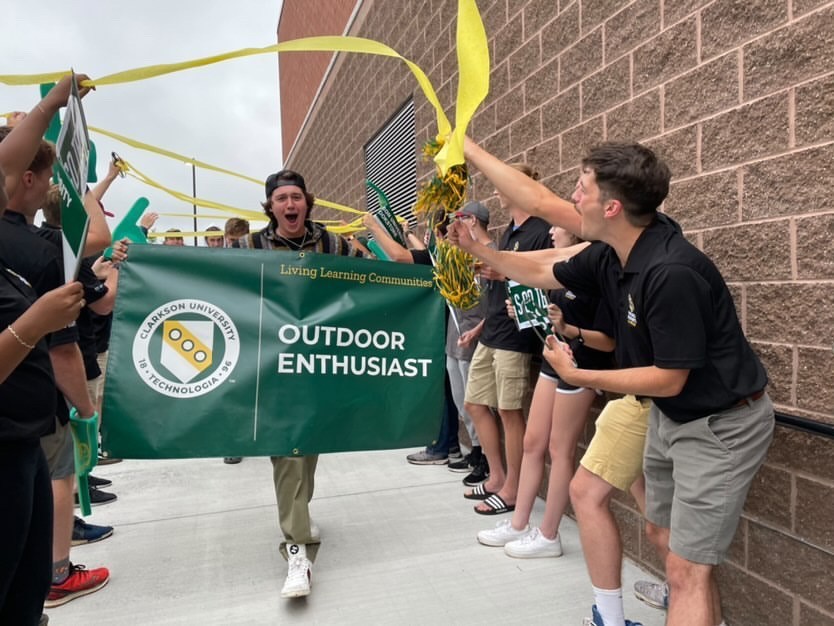 A student walking in a parade while holding a sign that says "Outdoor Enthusiast" with the Clarkson logo.