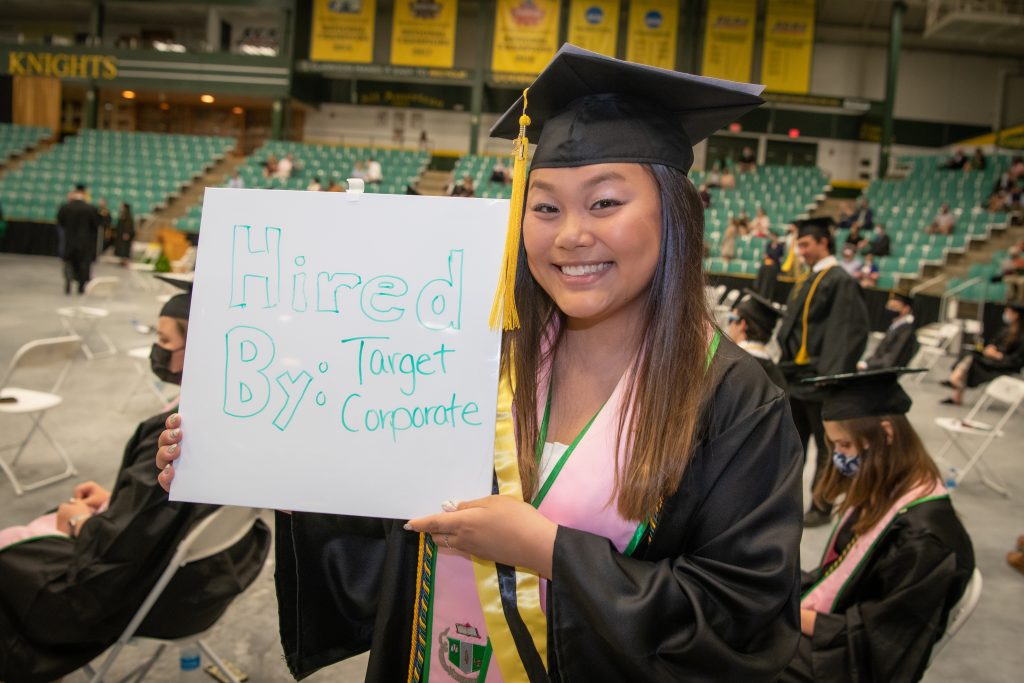 A women holding a whiteboard up with the words Hired By Target Corporate. The women is in her graduation cap and gown