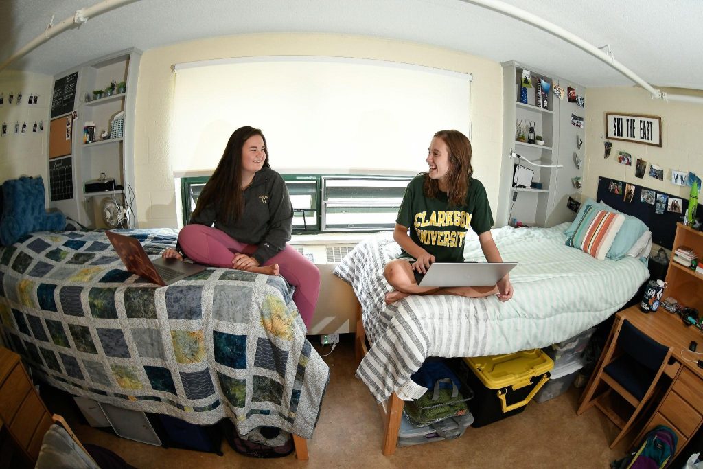 Two roommates sit on their respective dorm beds. They are looking at each other smiling.