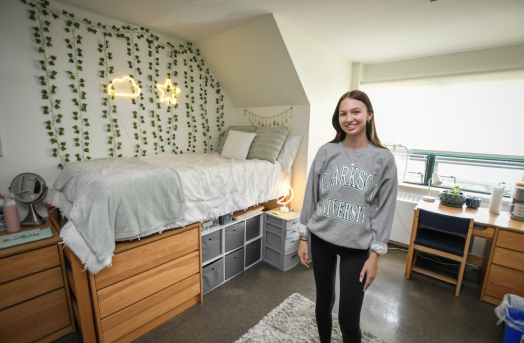 A student stands posed next to her bed in her dorm room after completing her decorating, which includes strings of small green decorations and lights shaped like a cloud and a star. 