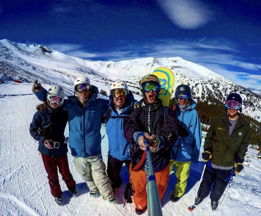 A group of 6 people taking a selfie with a selfie stick on top of a snow covered mountain 