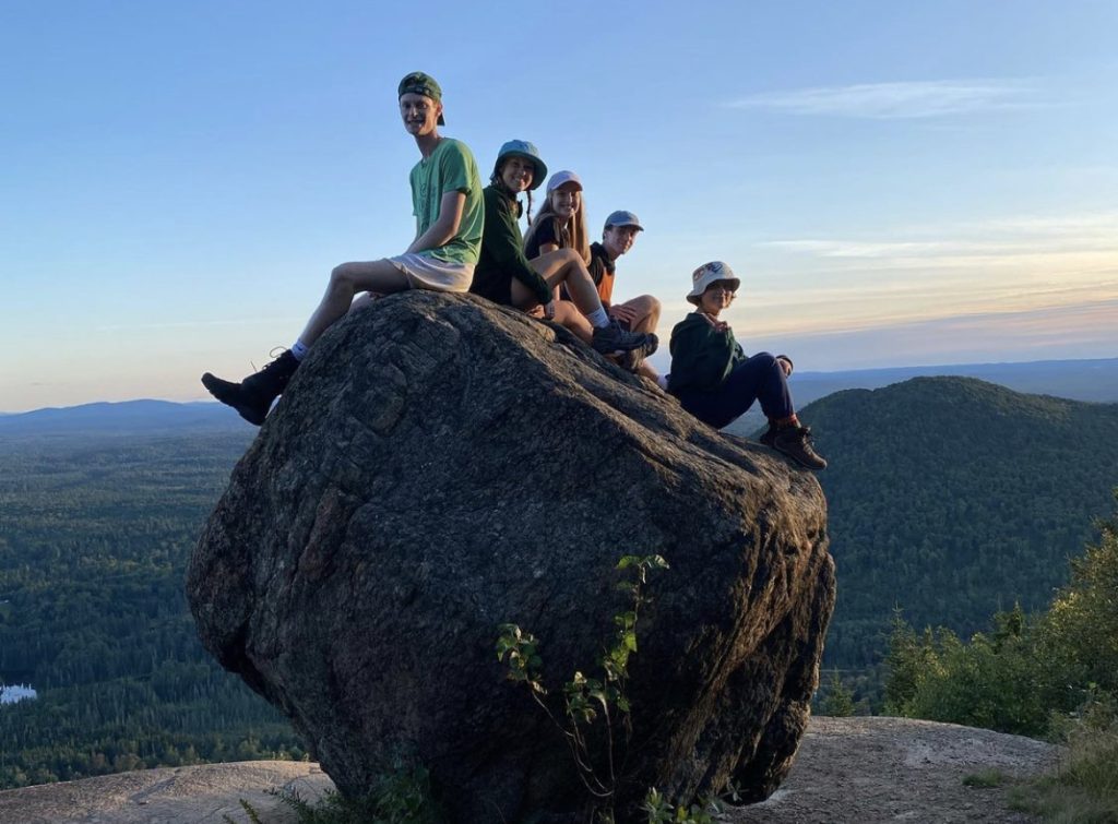 Five students sit smiling atop a large rock situated on the peak of a mountain with a cloud-wisped sky in the background