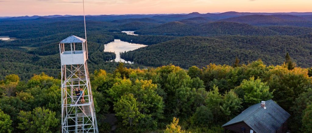 A fire tower on the left side of the photo overlooks a mountain-top view, complete with rolling hills and several bodies of water at sunset. 