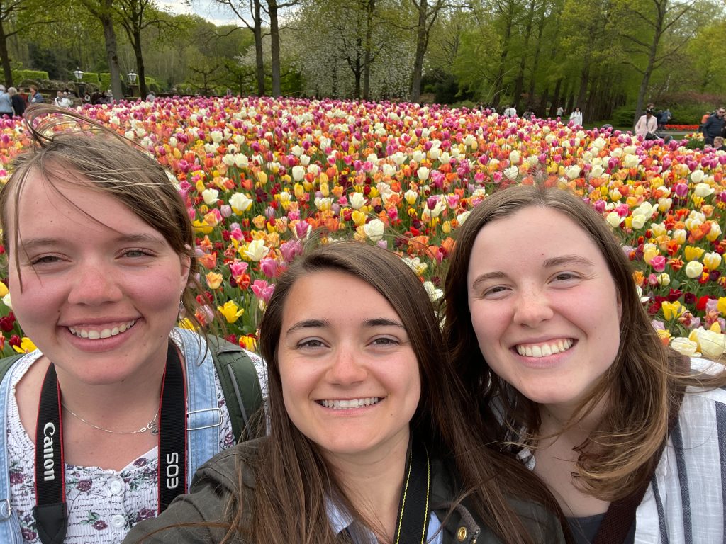 three people smiling into the camera in front of a color full bed of tulips with colors of yellow, pink, white, red