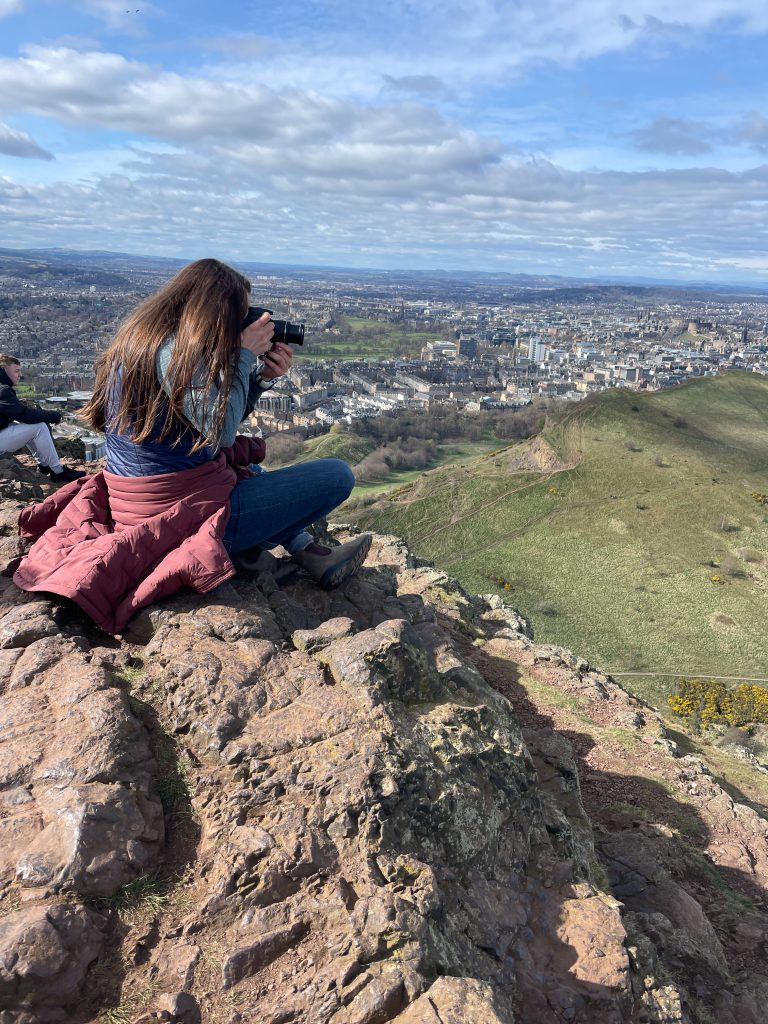 A person sitting on top of the mountain while taking a photo on a camera
