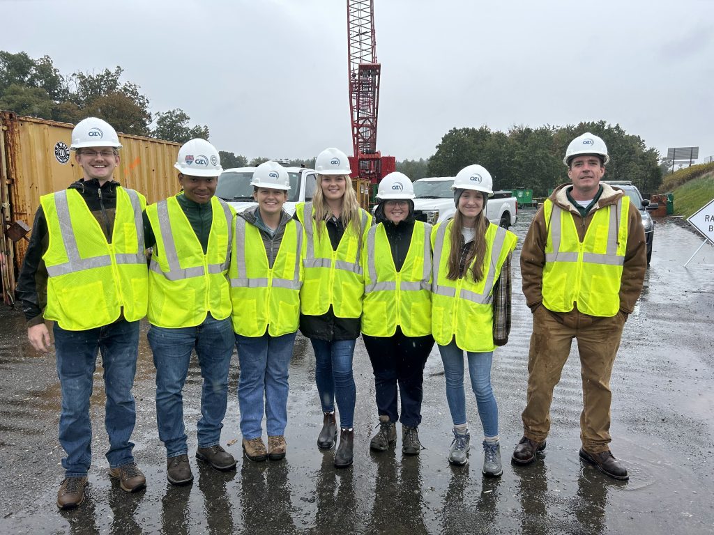 Image shows 6 Clarkson University Students an one professor standing shoulder to shoulder posing for a picture in their UV High Vis bright yellow construction vests. The group is standing in from on what seems to be a crane and some construction vehicles, including some white pick up trucks.