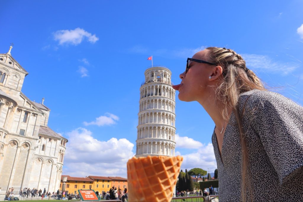 Carly sticking her tongue out to act as if she is licking the leaning tower of pisa with an ice cream cone at the edge of the image to have the tower as if its an ice cream